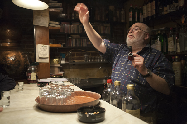 The owner of 't Dreupelkot jenever bar in Ghent behind the bar, with shelves of jenever bottles and a tray of small glasses